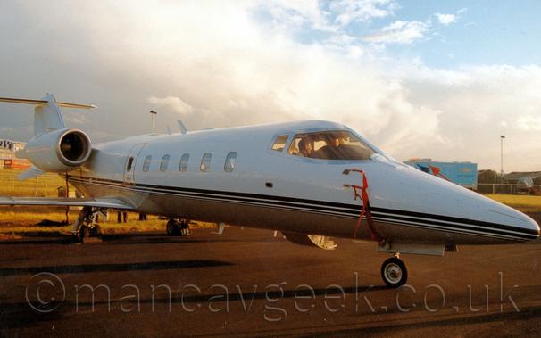 Side view of a sleek, white, twin engined bizjet parked facing to the right. The plane has a grey belly, and thin blue stripes running along the body. A couple of people are visible sitting in the planes cockpit, looking as if they are talking to each other. Bright sunlight coming from low on the right of the frame is casting long shadows to the left. In the background, blue sky gives way to thick, grey clouds.
