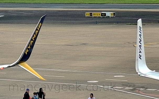 Closeup of the up-turned wingtips of 2 planes parked next to each other. The one on the right is the white inner face of a winglet, with blue "RyanAir" titles, which sits almost vertically on the end of the wing, while the one on the left is in 2 parts, a blue upper part with yellow "RyanAir" titles, and a yellow lower part, both parts canted out slightly. Grey concrete apron fills the space between and behind them.