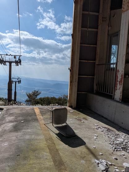Chair lying on its back casting a shadow near the entrance of the abandoned gondola station.  You can see the pylons for the cables dissappear down the mountain.