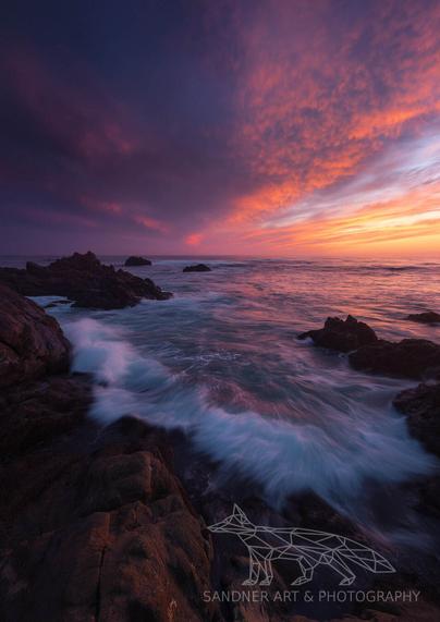 A vivid seascape taken during sunset at Phillip Island, with waves crashing against dark, jagged rocks in the foreground. The sky above burns with intense hues of magenta, orange, and violet, reflecting in the swirling water below. The dramatic light and motion create a powerful, moody atmosphere.