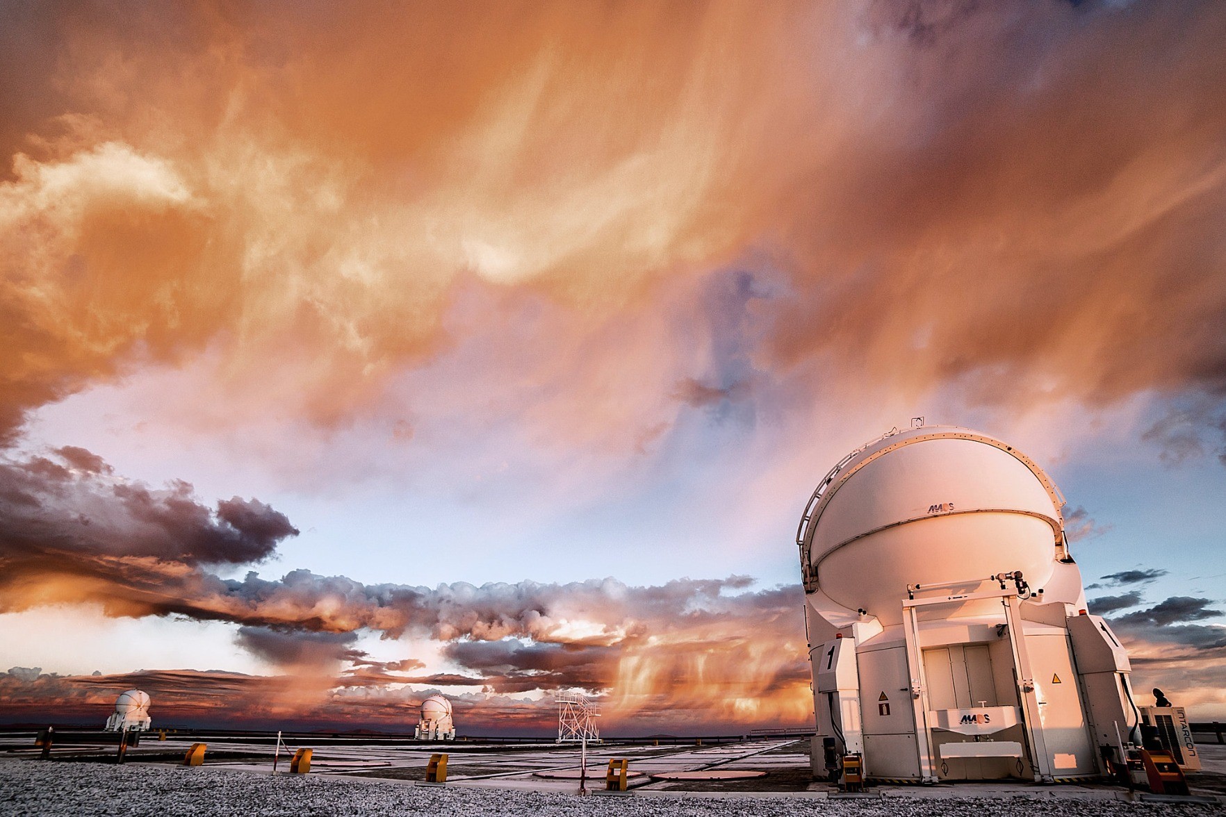 A stormy sunset over ESO's Paranal Observatory. There are three identical white telescope domes, one to the lower-right in the foreground and two to the background at the center left. The sky is blue, with several dark clouds. There are wispy veils of rain that don't quite touch the ground. These veils as well as the telescopes are bathed in the golden sunset light coming from the right (the Sun itself isn't visible in this image).