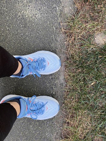 A patch of sidewalk next to a grassy verge, with a pair of lower legs & feet wearing brand new light blue Hoka trainers