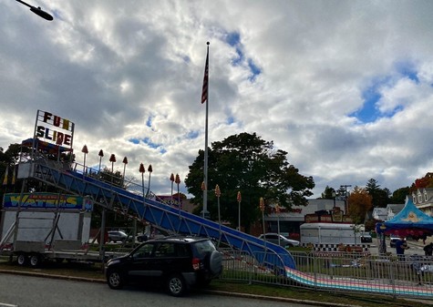 A photo of a fun fair being set up in the late afternoon, “FUN SLIDE” to the left, merry go round to the right, dramatic cloudy sky above, US flag just off center