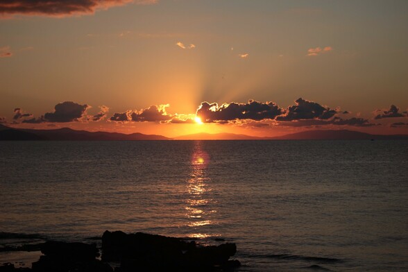 Sunrise over Greek island. Sun partly behind cloud, rays reaching over them, reflections on water. Slight lens flare.