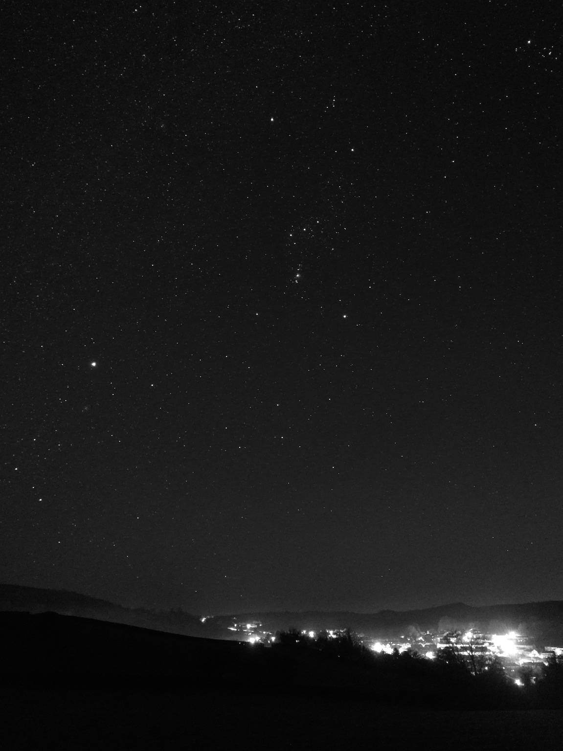 Monochrome night sky scene. View out into a valley, town lights on the right and a hill behind, mist in the middle distance on the left. In the far distance headlights from cars on a main road create an s shape. Near foreground completely dark fields with some trees creating silhouettes. Very starry sky, good transparency with stars right down to the horizon. Orion just right of centre with canis major in the lower left.  