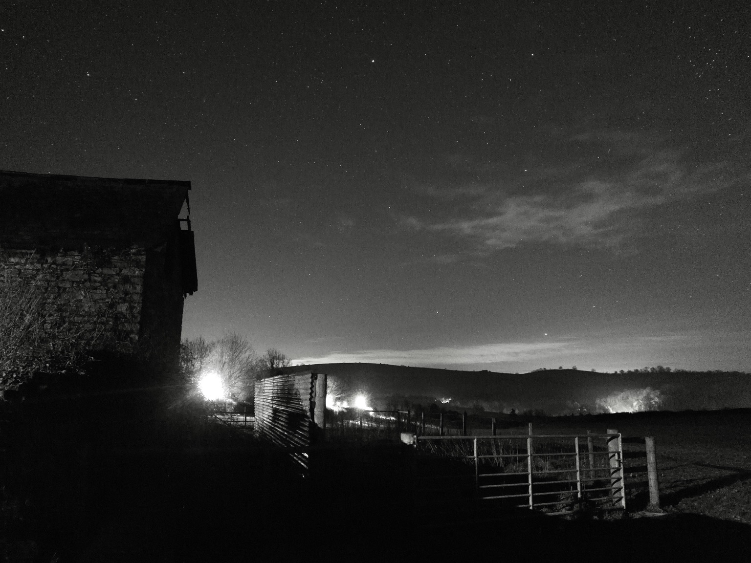 Monochrome night sky scene. Barn on the left with the stonework lit by a streetlight, a corrugated fence near the centre and a gate on the right. Streetlights in the middle distance are glaringly bright and a gritted lorry depot casts a light onto distant hills on the right. The sky is quite clear, although there’s a bank of cloud near the horizon and an area of high cloud towards the right, quite broken with tendrils. Many stars. Alkaid shines brightly at the top centre and a busy area of stars on the right is the ‘coma star cluster’ in Coma Berenices. Comet C/2022 E3 in Draco shows up as a pretty dim, fuzzy, smudge, with maybe some hint of a tail extending vertically but it is not clear