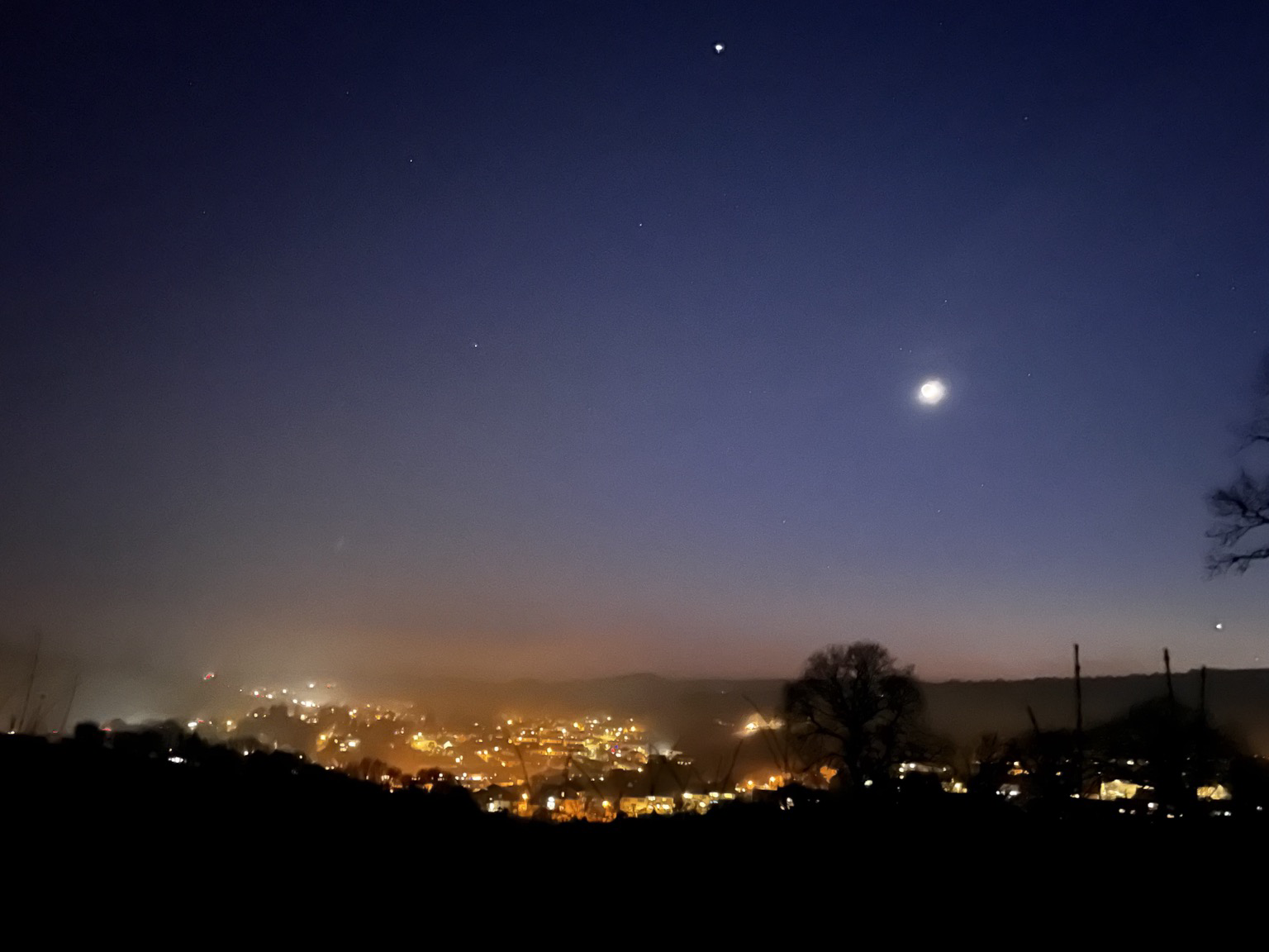 Scenic view of the town lights in a misty valley. Blue sky with a reddy orange area just above the distant hills. The waxing crescent moon is a bright blob, Venus a star like point close to setting in the lower right. Some background stars apparent elsewhere in the sky