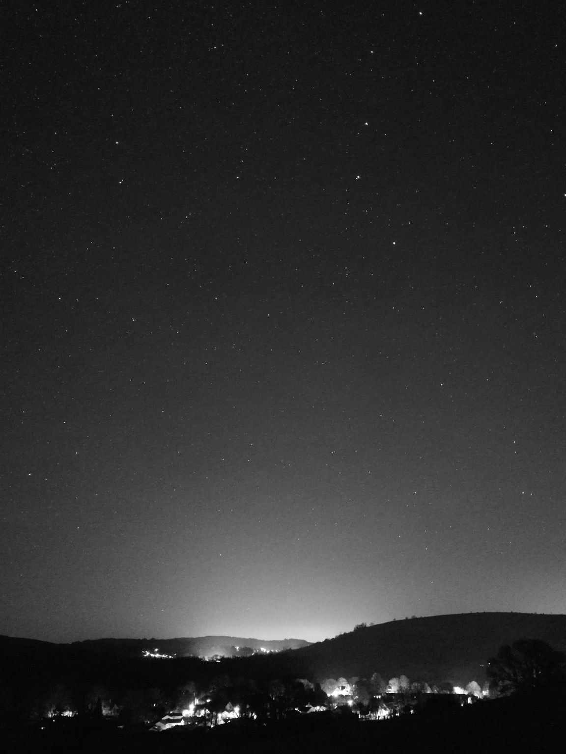 Monochrome night sky scene. Town lights in the middle distance, a large gentle hill on the right, and more distant lights beyond. Cloudless sky with many stars. The handle of the big dipper hangs down from the top right, and the ‘guardians of the pole’ are prominent in the top left. Comet C/2022 E3 is just to the lower right of the guardians, very faint and indistinct, just a tiny subtle smudgy point 