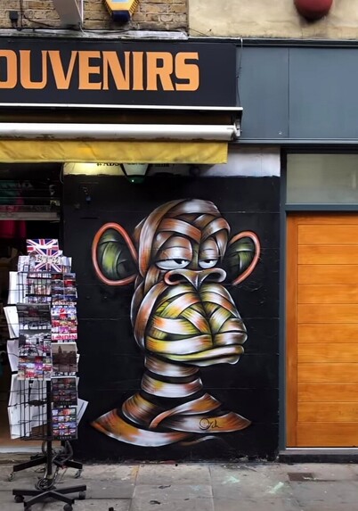 Streetartwall. On the 290x190m exterior wall of a souvenir store in London is sprayed a mural of a monkey with a bored look. The background is painted black. The monkey's head, the artist's trademark, is wrapped in colorful bandages. He looks like a colorful mummy in front of him.