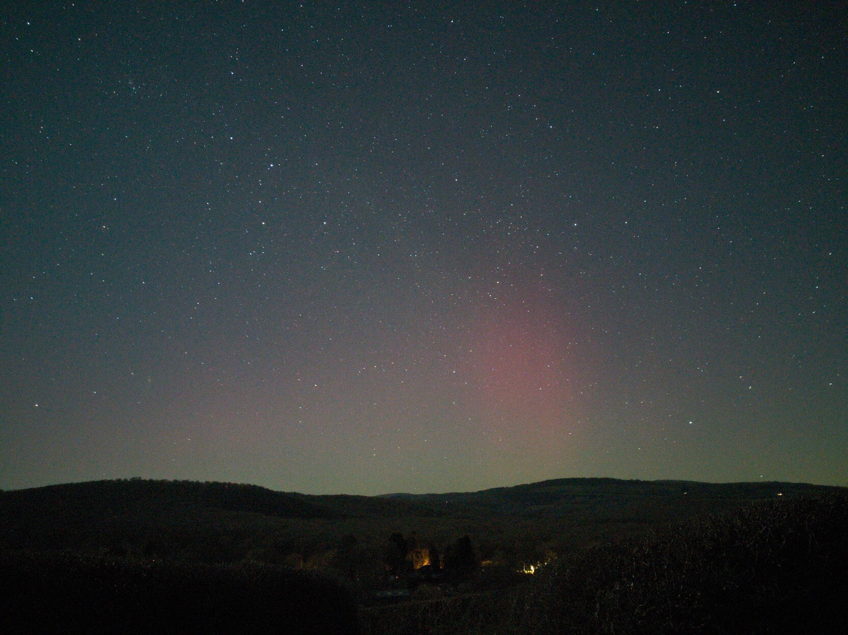 Nightscape. Lower area of the image is foreground of woods on gentle, rolling hills, a few lights in the mid-foreground from houses. The sky takes up the rest of the image, a cloudless starry sky, slightly blue from moonlight. A vague pink hue from aurora can be made out near the horizon, with a green layer lower down. In the centre right a brighter area of pink stands out.