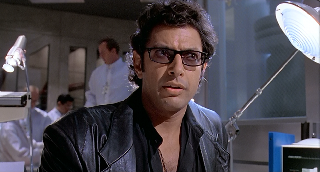 A screenshot of Jeff Goldblum as Dr. Ian Malcolm in Jurassic Park (1993) with a concerned look on his face, sitting in a lab with white-suited scientists behind him. Jeff may have been so preoccupied with whether he could play the part, he didn't stop and think if he should come back for the sequel.