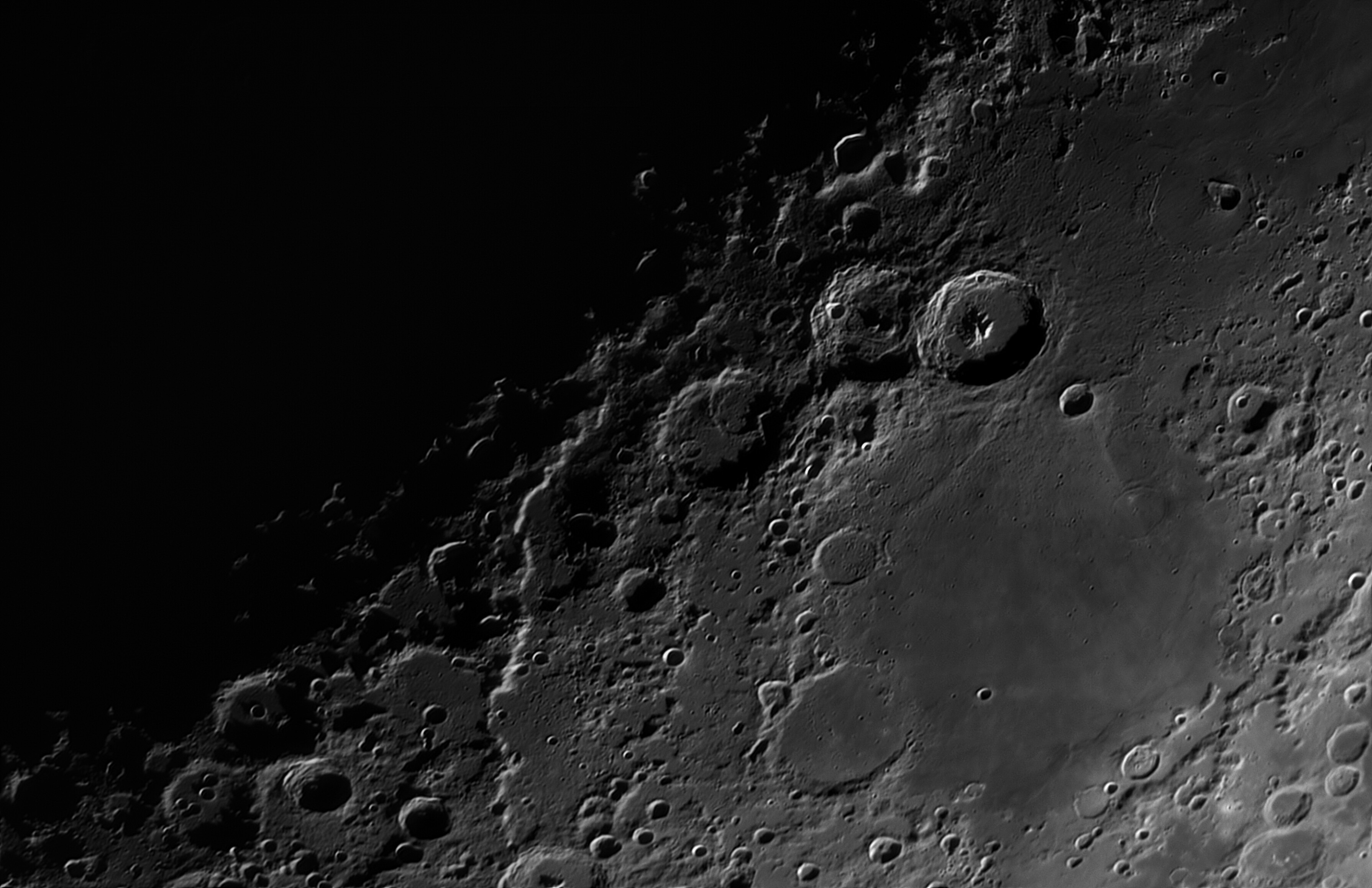 Monochrome closeup of the lunar surface. Mare Nectaris is a relatively plain area in the lower right. Above Nectaris are a series of large overlapping craters, shadows of the rims falling into the basins in the lower right of each one. Lunar escarpment Rupes Altai appears brights and winds its way from the centre down towards the bottom of the frame. The top left area is in shadow appearing completely dark. 