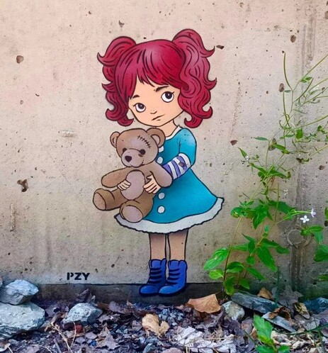 Streetartwall. On a beige concrete wall is sprayed/painted a little comic mural of a little girl with her teddy bear. She has big eyes, a small snub nose and mouth, two red braids and wears a light blue short dress with white hem, blue boots underneath. She holds the brown teddy bear sideways in front of her. There are gray stones in front of the mural and a green plant with white flowers climbing up the side. Cute.