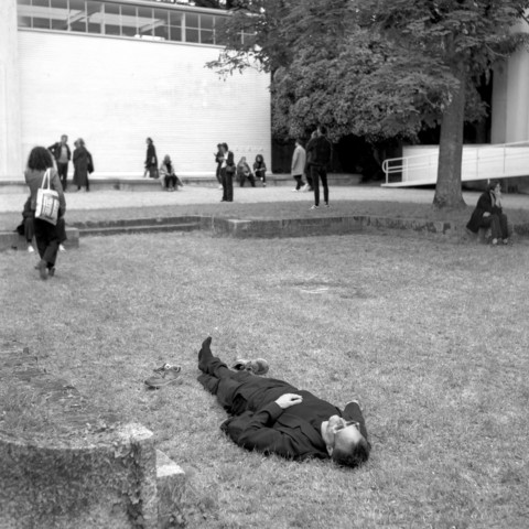 A man laying with his shoes off on the lawn in front of the Austrian pavillion at the Architecture Biennale in Venice. People strolling in the background.