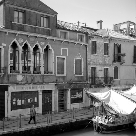 A window view from a house at the end of the Via Garibaldi in Venice on the opposite side of the canal, with a vegetable market on a boat, house facades and a man walking along it.