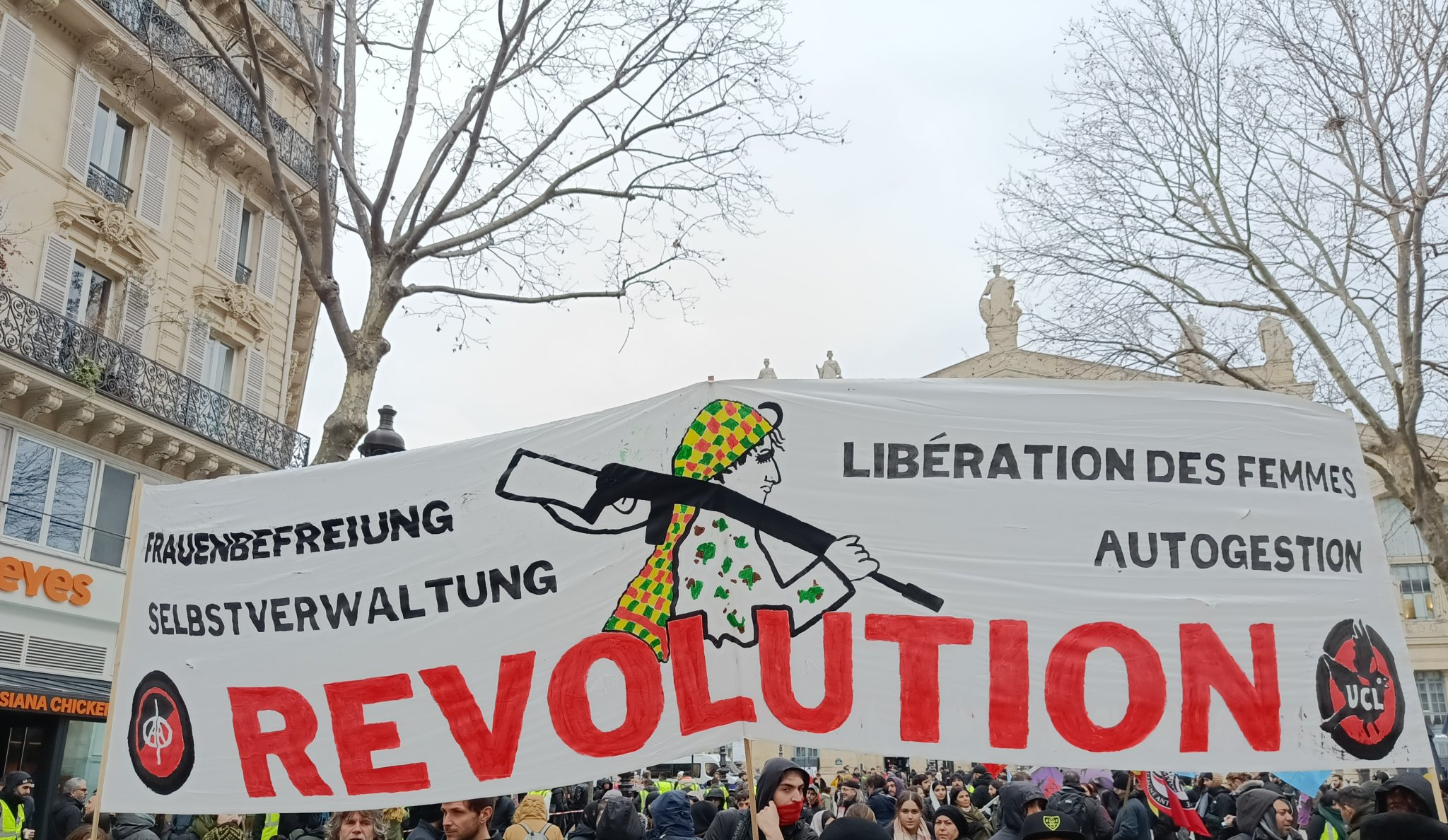 Banderole bilingue Die Plattform UCL : Libération des femmes, Autogestion avec une femme kurde armée entre le texte en allemand et en français et en dessous en grosses majuscules rouges Révolution avec logo Die Plattform à gauche et UCL à droite