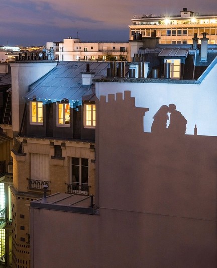 Photography. A color photo of a view over the rooftops of Paris with a shadow play on a house wall. The color photograph was taken in the evening. A view of houses opposite, whose windows are partially illuminated. The evening sky is slightly cloudy. Blue-grey light falls on the blank white wall of the house opposite. Next to the shadow of a roof with four small chimneys, a woman and a man can be seen kissing. The shadow of a wine bottle can be seen behind them.