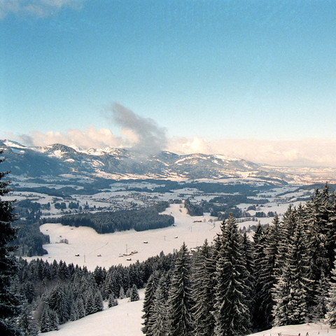 Panoramablick auf eine alpine Winterlandschaft. Im Vordergrund dominieren schneebedeckte Tannen, welche die linke Seite des Bildes einrahmen. Darüber hinaus erstreckt sich ein offenes, hügeliges Terrain mit Feldern und verstreuten Bäumen, die ebenfalls von Schnee überzogen sind. Ein Dorf ist in der Ferne erkennbar, eingebettet in das Tal zwischen den Hügeln. Im Hintergrund erheben sich weitere schneebedeckte Berge, die teilweise von Wolken umgeben sind. Der Himmel ist größtenteils klar mit eine…