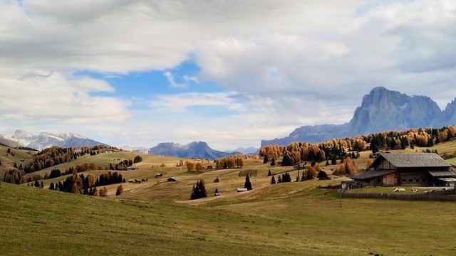 Panoramic photography - Rolling hills with autumn-colored trees, a few small cabins, and a mountain range in the background under a partly cloudy sky. 