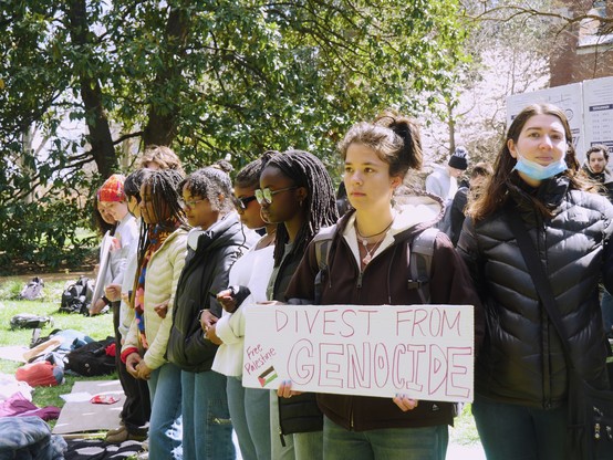 A line of students, including those of visible African and European descent stand in protest with their arms linked. One holds a sign reading "Divest from Genocide / Free Palestine"