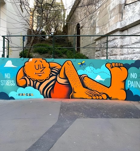 Streetartwall. The mural of a friendly man in the clouds was sprayed/painted on a long, low street wall. An orange man is lying feet first between blue clouds and a flying butterfly. He is wearing a striped T-shirt, red shorts and his head is shaped like a sun. He has his eyes closed and is enjoying his time off. Next to the figure is written on the left: "No stress" and on the right "No pain". (Behind the mural is a hill with a tree and green hedges, between two houses)
