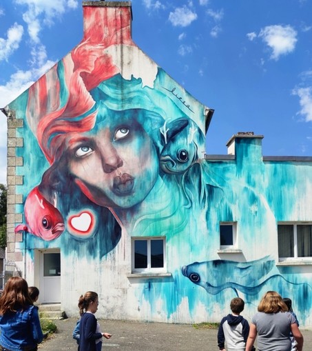 Streetartwall. A mural of a young woman surrounded by fish has been sprayed/painted on the low exterior wall of a two-storey brick building. The beautiful woman is painted in shades of red and turquoise, and her radiant face looks up into the blue sky. She makes a kissing mouth. Around her, green and red fish swim through the mist of color with an Asian painting style. A white heart with a red border completes the mural. (In the photo, some passers-by are standing in front of the wall to admire it).
Info: The mural is intended to express a possible rapprochement between humanity and the world of animals in an ecologically responsible way and is based on a Japanese fable by Miyasaki.
