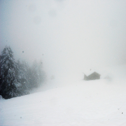 Eine kleine Hütte, teilweise vom Nebel verhüllt, in einer verschneiten Landschaft. Schneeflocken fallen, sichtbar vor dem dunklen Hintergrund der Hütte und Bäume. Die Sicht ist durch den dichten Nebel stark eingeschränkt, was für eine ruhige, isolierte Stimmung sorgt.