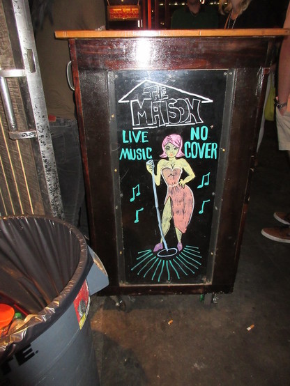 Erasable sign board. 
Text at top "THE MAISON"
"LIVE MUSIC"
"NO COVER"

Cartoon artwork depicts a woman in a slinky dress holding a microphone, surrounded by musical notes. 