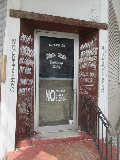 Glass doorway in corner wooden building. 
Lettering  applied to door. 
At top, address" "1940 Eagle St."
Larger letters in playful font, name of bar: "Sista Sista"
"Bar & Lounge - 30 and up"

Lower part of door, severe font: "NO Drugs - NO shirt shoes service"

Matching signs on either side of the doorway,  weathered paint, white letters on red background on wood: "NO! DRUGS ALLOWED AT ALL *
 THANK YOU OWNER  * 
NO! DRUGS WILL CALL POLICE"

Matching text is on wooden support columns on either s…