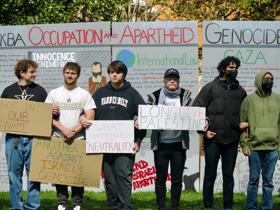 Six protesters with linked arms stand in from of a wall explaining and denouncing "Nakba; Occupation and Apartheid; Genocide." Several of them wear Vanderbilt shits and hold signs reading "Not In Our Name", "Another Jew Against Israel's Genocide", "What Happened to Neutrality", "Long Live Palestine"