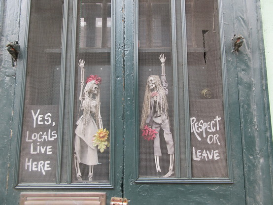 Exterior view of screened windows in weathered wooden frame. Hanging inside are a pair of skeleton dolls in bride and groom costumes holding flowers.  To either side are hand lettered signs. 
At left, text: "Yes, Locals Live Here"
At right, text: "Respect or Leave"