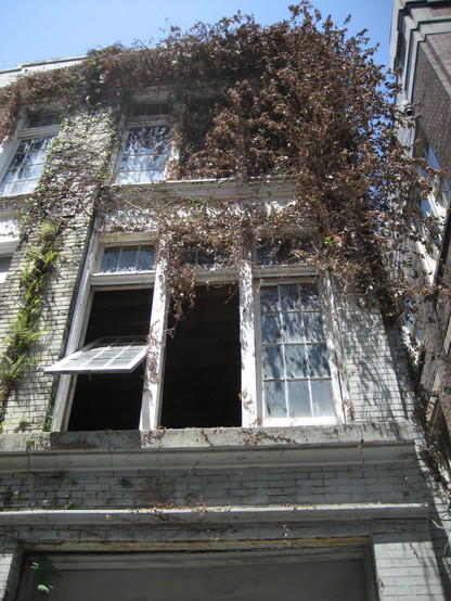 Exterior view towards upstairs of 3 story brick building.  It is partly covered with vines and plants.  Windows and shutters on the middle floor are open or missing, giving appearance that at least that part of the building is vacant or abandoned.