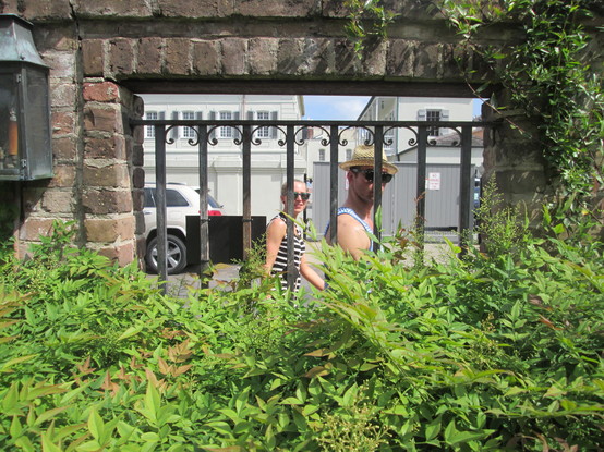 View from interior garden, with a barred window in brick wall.  Lush plants are seen at bottom and right of photo. Through the window a young woman and a young man, both in sunglasses and casual clothes, are seen walking on the sidewalk; they turn to their heads to look in the window towards the photographer.  In background 2 story colonial era buildings (part of the old Ursulines Convent) are visible.