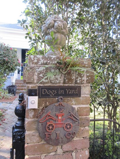 Brick rectangular pillar besides the gate to a house. It has a sculpture of a bowl of fruit on top. An old metal fire insurance plaque is attached to the pillar, with a design of a 19th century hand pump fire engine over the letters "F.I. Co."    Two buttons for doorbells are attached to left. Above the plaque is a sign reading "Dogs in Yard". 