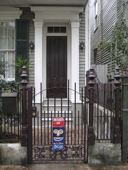 Metal gate in front of stairs leading to doorway of wooden 19th century neo-classical style house. Hand drawn sign is on the gate. In center is a cartoon drawing of a face, 2 round eyes, small nose, and a wide grinning mouth with sharp teeth. 
Above the face is lettering "BEWARE OF".
Below is text "Alligator", with the word alligator crossed out.
At bottom is text "Dogs". 