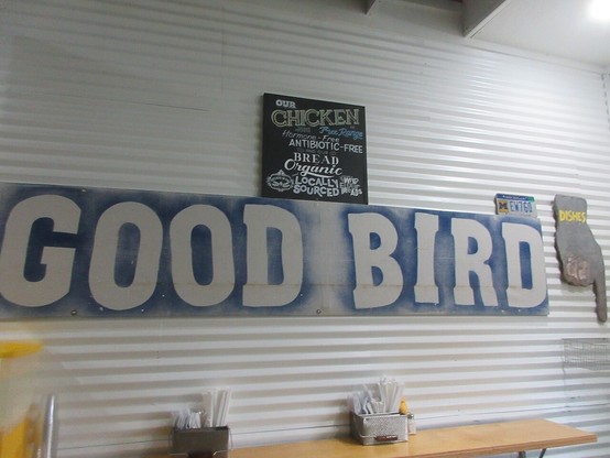 Interior of small restaurant. Large sign on wall reading: "GOOD BIRD". 
A smaller sign at top reads: "Our CHICKEN is Free Range - Hormone-Free Antibiotic-Free - and our Bread is Organic Locally Sourced Wild Flour Bread".  

At bottom of photo is a restaurant counter with containers holding paper napkins, straws, salt and pepper shakers. 