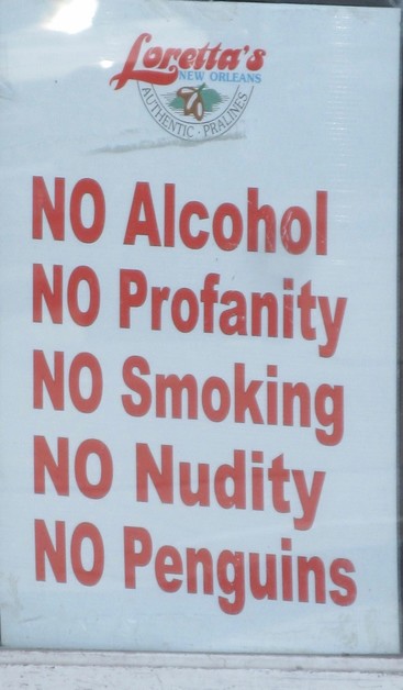 Sign, red lettering on white. 
At top "Loretta's New Orleans Authentic Pralines" with logo depicting 3 pecan nuts and a leaf.
Larger text below: "NO Alcohol - NO Profanity - NO Smoking - NO Nudity - NO Penguins". 
