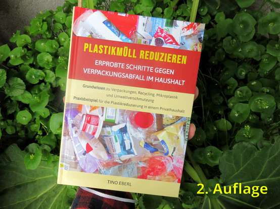 Ein Buch mit dem Titel "PLASTIKMÜLL REDUZIEREN - ERPROBTE SCHRITTE GEGEN VERPACKUNGSABFALL IM HAUSHALT" von Tino Eberl wird gehalten. Das Cover zeigt eine Sammlung von Plastikabfällen, wie Flaschen und Verpackungen, um das Thema Müllreduzierung zu illustrieren. Das Buch gibt  praktische Tipps und Grundwissen über Recycling und Umweltverschmutzung zu bieten. Es ist vor einem Hintergrund von grünen Pflanzen fotografiert.