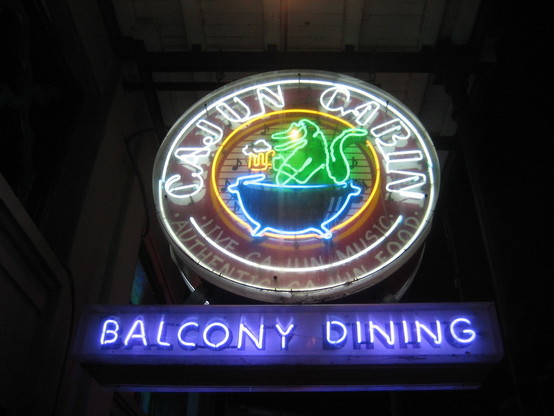Night view of illuminated circular neon sign. 

Text around outer ring: "CAJUN CABIN - Live Cajun Music - Authentic Cajun Food".  

Cartoon artwork in center depicts an alligator sitting in a large pot, holding a mug of beer.

Rectangular neon sign below: "BALCONY DINING".
