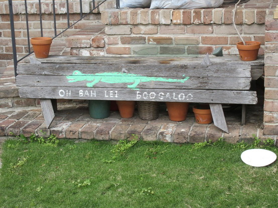 Wooden bench beside brick staircase. Clay plant pots are on either end of the bench, with more pots underneath.  Along the length of the bench is painted a green alligator. 
Text underneath: "OH BAH LEI BOOGALOO"