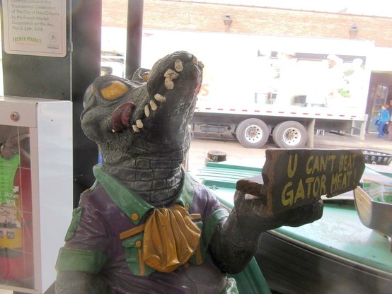 Multicolored statue of an alligator standing upright, licking its lips and rolling its eyes up, wearing purple green and gold colored human clothes. One hand is raised holding a tray.  On the tray is a wooden sign reading "U CAN'T BEAT GATOR MEAT!"