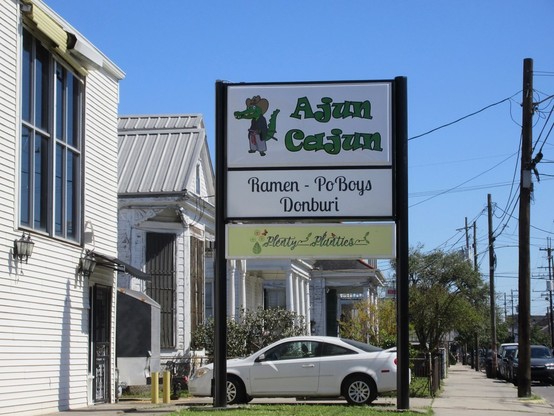 Street with row of 1 and 2 story buildings. 
Large business sign, painted glass in metal supports. 

To sign text: "Ajun Cajun". 
Cartoon of standing anthropomorphic alligator, wearing a cowboy hat and with a belt with a samurai style sword.

Lower sign text: "Ramen - Po' Boys - Donburi"
