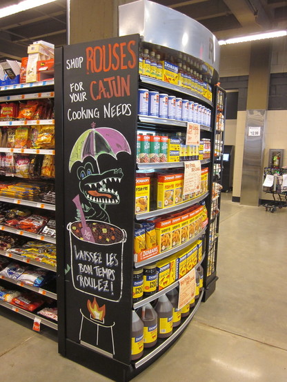 Interior of a supermarket. Shelf with local spices and cooking supplies has large chalkboard sign. 

Text: "Shop ROUSES For Your CAJUN Cooking Needs". 

Multicolor chalk cartoon of an alligator stirring a large cooking pot.  A colorful parasol is above the gator's head; the gator smiles broadly with rows of sharp teeth. 
Text on the cooking pot: "Laissez les Bon Temps Roulez!"  (Louisiana French for "Let the Good Times Roll".) 