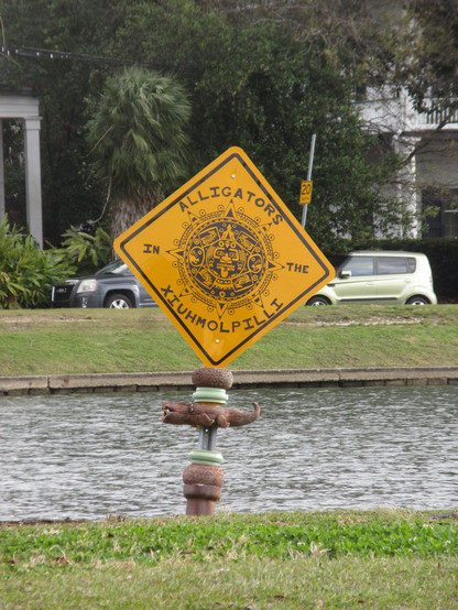 Bayou body of water with grassy banks, houses seen in background on far shore. In center is a square metal sign, black text and artwork on bright yellow.  Text: "ALLIGATORS IN THE XIUHMOPOILLI".  The text surrounds round artwork, copied from the center of the famous Aztec Sun Stone, depicting a stylized human face with tongue sticking out surrounded with Aztec glyphs in the form of reptile heads and an outer circle with points sticking out, a mythological depiction of the Sun. 
The pole holding…