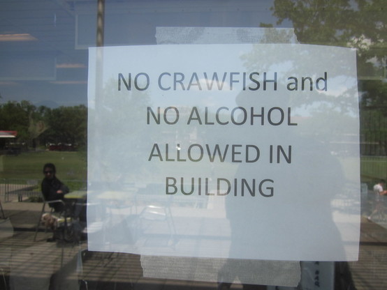 Printed paper sign taped in glass door window.  A wide yard with outdoor tables seen in reflection. 

Text: "NO CRAWFISH and NO ALCOHOL ALLOWED IN BUILDING"

