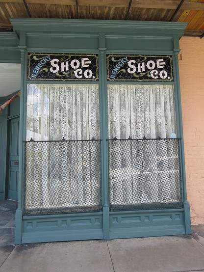 Shop window in wooden frame, sticking out into sidewalk under a covered gallery, 1890s/1900s style.  Upper part of twin windows has stained glass with art nouveau style lettering reading "RENECKY SHOE CO.".  Lower part of windows is clear, with white curtains inside and metal mesh barrier. 

