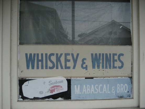 Old neighborhood grocery shop window with painting, style c. 1930s - 1950s.  Text: "WHISKEY & WINES". 
Below, artwork depicting a load of bread wrapped in paper, with text: "Sunbeam Batter Whipped - COMPARE". 
At bottom right, text: "M. ABASCAL & BRO". 