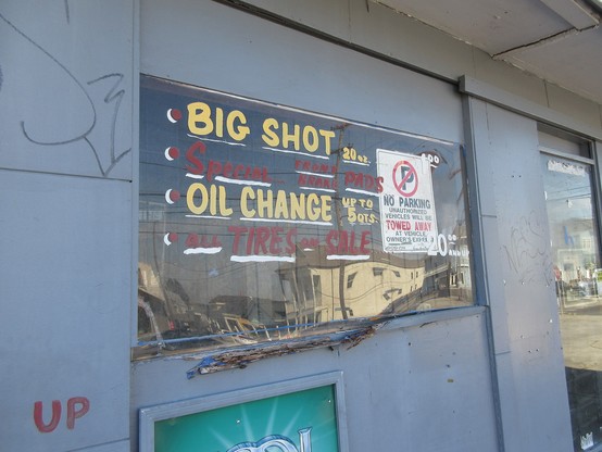 Auto service shop window with hand painted lettering.
Text: '* BIG SHOT  20 oz - 
* Special Front Brake Pads - 
* OIL CHANGE up to 5 qts - 
* All TIRES on SALE".   

A "No Parking" sign is affixed to the window.
