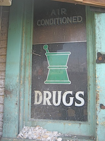 c. mid-20th century shop window in wooden frame, pile of old plaster rubble seen at bottom.   Text on upper part of window: "AIR CONDITIONED".  At center, stylized artwork depicting a mortar and pestle.  Text below: "DRUGS". 
