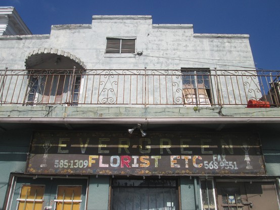Faded 2 story shop front with balcony upstairs hanging from which is a faded wooden sign, brown background painted with letters in random alternating colors.  

Text: "EVERGREEN FLORIST ETC".  On either side are matching artwork depicting a vase in the form of a woman carrying a basket on her head, at left with a 7 digit telphone number, and another number at right labled "FAX". 
